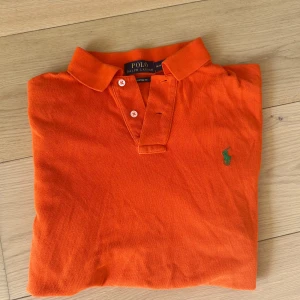 Orange pikétröja från Ralph Lauren - Snygg orange pikétröja från Polo Ralph Lauren i bomull, custom fit. Klassisk krage, knäppning med två knappar och grönt broderat logomärke på bröstet. Perfekt för dig som vill sticka ut med färg och stil.