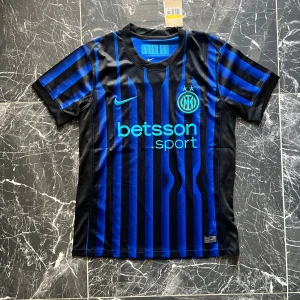 Inter Milan randig fotbollströja Nike - Snygg Inter Milan fotbollströja från Nike med klassiska blå och svarta ränder. Kortärmad modell med klubbmärke och sponsortryck på bröstet. Tillverkad i lätt och ventilerande polyester, perfekt för match eller träning. Modern passform och sportig look.