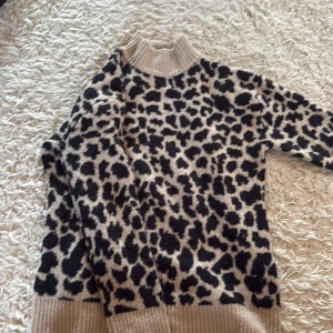 Leopardmönstrad polotröja från NA-KD - Säljer en beige och svart polotröja från NA-KD med leopardmönster. Tröjan har ribbad polokrage och muddar, är långärmad och stickad i mjukt material. Perfekt för dig som vill sticka ut med ett djuriskt print och hålla dig varm under kyliga dagar.