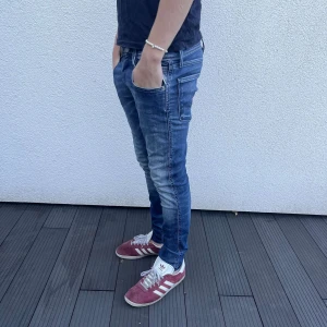 Replay Anbass jeans - Snygga blå Replay Anbass jeans med skinny passform och coola slitningar. Klassisk femficksdesign, tydliga sömmar och läderpatch bak i midjan. Jeansen är gjorda i stretchigt denim för skön komfort och sitter tajt längs benen.