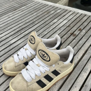 Beige Adidas Campus sneakers mocka - Adidas Campus sneakers i beige mocka med svarta detaljer och klassiska tre ränder på sidan. Skorna har vit platt sula, rund tå och vita snören. Adidas logga på tungan och hälen. Perfekt för skolstart!