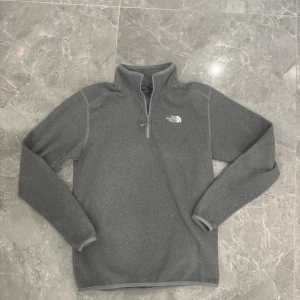 Grå fleece-tröja från The North Face - Säljer en grå fleece-tröja från The North Face i storlek S. Tröjan har half zip framtill, hög krage och långärmade ärmar. Perfekt för kyliga dagar och outdoor-häng. Mjuk och skön polyester som håller värmen. Diskret logga broderad på bröstet.