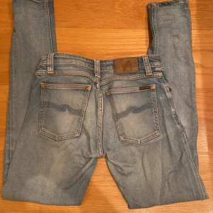 Ljusblå skinny jeans från Nudie - Säljer ett par ljusblå skinny jeans från Nudie med slitna detaljer och hål på knäna. Storlek w27/L32 Klassisk femficksmodell med Nudie-logga på bakfickan och snygga kontrastsömmar. Fet färg med sköna slitningar 
