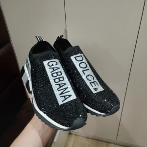 Svarta sneakers Dolce & Gabbana - Säljer ett par svarta sneakers från Dolce & Gabbana med glittriga paljetter över hela ovandelen och tydliga logotexter. Skorna har en chunky, vit och svart sula med grå detaljer och ribbad undersida. Stretchig ovandel och slip-on modell för skön passform.