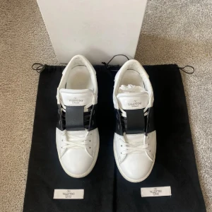 Valentino Garavani Open sneakers - Snygga Valentino Garavani Open sneakers i vitt skinn med bred svart läderdetalj över mitten. Klassisk rund tå, platt sula och coola nitar på hälen. Skorna har snörning och en stilren design som passar perfekt till streetwear och trendiga outfits.