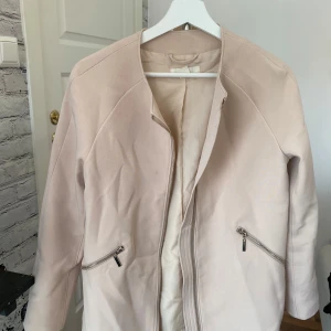 Beige kappa från H&M med dragkedja - Snygg beige kappa från H&M i storlek 40. Jackan har rund halsringning, två snedställda fickor med silverfärgade dragkedjor och stängs med dragkedja framtill. Materialet känns mjukt och har en stilren look som passar till många outfits.