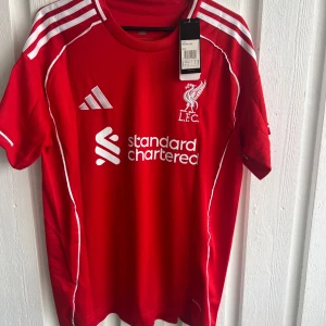 Liverpool röd matchtröja Adidas - Liverpool FCs officiella röda matchtröja från Adidas med vita detaljer och tryck. Kortärmad med klassiska vita ränder på axlarna, klubbmärke och sponsorlogga på bröstet. Namn och nummer Frimpong 30 på ryggen samt UEFA Foundation-märke på ärmen.