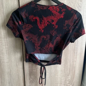 Svart croppad topp med röda drakar - Cool croppad topp från SHEIN med korta ärmar och ett rött drakmönster över hela. Toppens baksida har snörning nedtill som ger en snygg detalj och sitter tajt mot kroppen.