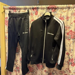 Svart trackjacka från Palm Angels L - Palm Angels svart trackjacka med vita ränder längs ärmarna och dragkedja framtill. Jackan har ståkrage och logotypen broderad på bröstet. Materialet är en mix av polyester och bomull, vilket ger en mjuk och sportig känsla. Perfekt för en streetwear-look.