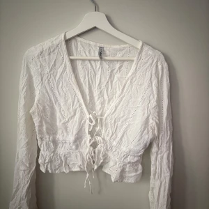 Vit croppad blus med snörning - Supersöt vit blus med croppad passform och långa ärmar. Blusen har snygg snörning framtill, v-ringad hals och volangdetaljer vid midjan. Materialet är lätt och bomulligt med broderade hål för extra vibe.