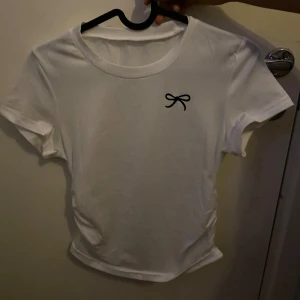 Vit t-shirt med svart rosett - Vit t-shirt med korta ärmar och rund hals. På bröstet finns en liten svart rosett som detalj. Modellen är enkel och har en något figurnära passform. Perfekt för dig som gillar stilrena plagg med en söt twist. Materialet känns mjukt och skönt, troligen bomull. Använd en gång bara 🩵