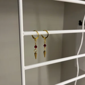 Guldiga örhängen med röd detalj - I rostfritt stål och handgjort. Snygga guldiga örhängen med små röda pärlor och spetsiga hängen. Perfekt för dig som vill ha en edgy och cool stil. De har en rund form upptill och hängande konformad detalj som ger extra attityd.