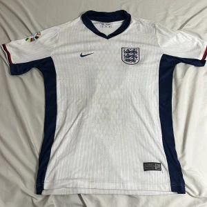 England Bellingham 10 fotbollströja L - Snygg England fotbollströja med Bellingham och nummer 10 på ryggen. Tröjan är vit med marinblå detaljer längs sidorna och kragen, samt röd och blå rand på ärmslut. Broderat England-emblem på bröstet och Nike-logga. Tillverkad i lätt och ventilerande material.