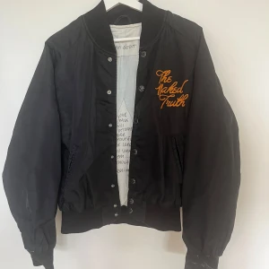 Svart bomberjacka från Nudie Jeans - Svart bomberjacka från Nudie Jeans med orange broderad text 'The Naked Truth' på bröstet. Jackan har ribbade muddar, knappstängning och innerfoder med handskriven print. Snygg, klassisk passform och coola detaljer som sticker ut. Finns en liten fläck på innefodret, säljer den därför billigt.