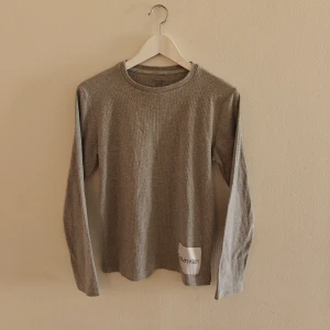 Calvin Klein tröja - Snygg grå långärmad tröja från Calvin Klein med diskret ribbad struktur och rund hals. Tröjan har en liten vit logotyppatch nedtill och är gjord i mjuk bomull med lite stretch för skön passform. Perfekt för dig som gillar stilrena och enkla plagg.