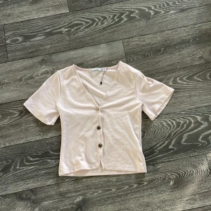 Ljusrosa v-ringad topp från 157 - Säljer en ljusrosa topp från 157 i storlek XS. Den har v-ringning, korta ärmar och tre snygga knappar framtill. Modellen är croppad och gjord i mjuk bomull, perfekt för dig som gillar enkel och fräsch stil.