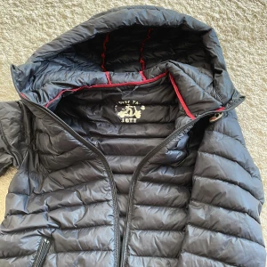 Svart dunjacka från JOTT med huva - Snygg svart dunjacka från JOTT med huva och röda detaljer på insidan. Jackan har quiltad design, två fickor med dragkedja och ett broderat JOTT-märke på ärmen. Perfekt för kalla dagar och har en stilren look som funkar till det mesta. Har även använts av kille i storlek xs o funkar perfekt