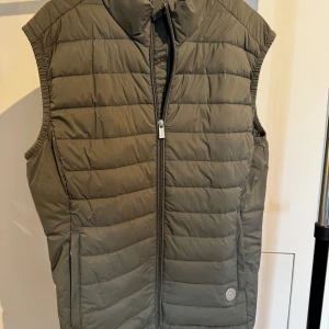 Olivgrön dunväst från Snoot Stockholm - Olivgrön dunväst från Snoot med quiltad design och hög krage. Västen har dragkedja framtill, två sidofickor med logga i nacken.