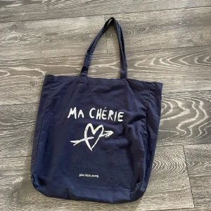 Mörkblå shoppingväska Gina Tricot Young - Stor och rymlig mörkblå shoppingväska från Gina Tricot Young med trycket 'MA CHÉRIE' och ett hjärta med pil på framsidan. Väskan har två långa handtag och är tillverkad i bomull. Perfekt för att bära med sig allt man behöver under dagen.