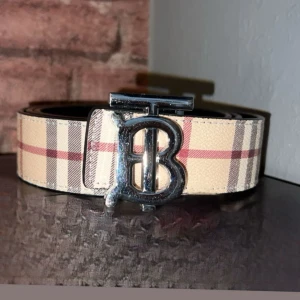 Burberry bälte med B-spänne.       125cm - Säljer ett snyggt Burberry bälten med ikoniskt B-spänne i silver. Ena sidan av bältet har klassiskt rutigt mönster i beige, rött och grått, medan det andra är helsvart med subtilt mönster. Är tillverkade i skinn och har en bred, stilren form som ger en lyxig vibe.