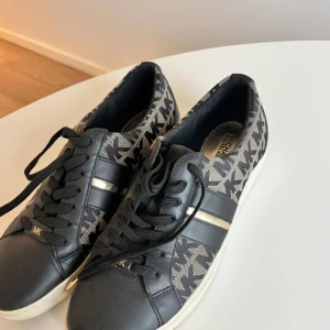 Svarta sneakers från Michael Kors - Snygga svarta sneakers från Michael Kors med MK-logga i mönster på sidan och detaljer i silver. Skorna har snörning och en vit platt sula. Materialet är en mix av skinn och textil, vilket ger en cool kontrast. Perfekta för dig som gillar stilrena och trendiga skor.