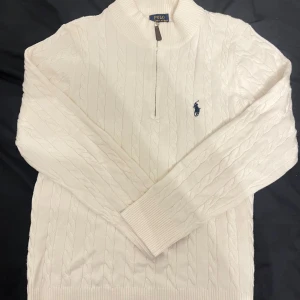 Vit kabelstickad tröja Polo Ralph Lauren - Snygg vit kabelstickad tröja från Polo Ralph Lauren med half zip och broderad marinblå logga på bröstet. Tröjan har långärmad design och ribbade muddar vid ärmslut och nederkant. Perfekt för dig som gillar klassisk stil.