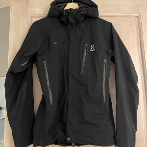 Svart vindjacka från Haglöfs - Snygg svart vindjacka från Haglöfs med huva och flera dragkedje fickor. Jackan är i mycket gott skick! Storlek S, pris 1200:-
