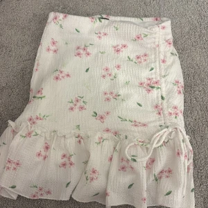 Blommig volangkjol från Gina Tricot - Supersöt kort kjol från Gina Tricot i storlek XXS. Kjolen är vit med rosa blommor och gröna blad, har volangkant och knyt i sidan. Materialet är lätt och krispigt, perfekt för varma dagar. Passformen är normal och kjolen har en omlott-look.