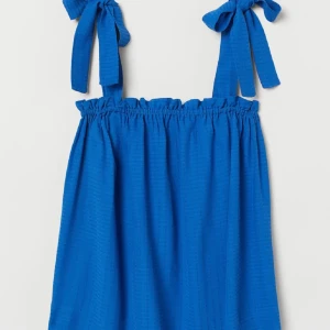 Blå topp med knytband - Supersöt blått linne med knytband på axlarna och rynkad kant upptill. Kort modell som är perfekt för varma dagar. Materialet har en lätt struktur som ger en cool vibe. Enkel och stilren design som är lätt att matcha med sneakers eller sandaler.