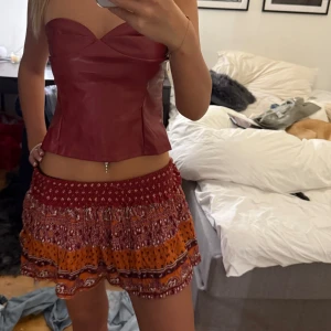 Röd och orange kortkjol från Superdry - Supersöt kortkjol från Superdry i röd och orange med blommigt mönster och volangkant. Kjolen har smockad midja som sitter skönt och ger en loose passform. Perfekt för sommarens alla dagar, lätt och luftig i bomull. 