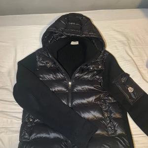 Svart pufferjacka från Moncler - Svart pufferjacka från Moncler med stickade ärmar och huva. Jackan har quiltad front i blankt material, dragkedja och en snygg ficka med Moncler-logga på ärmen. Perfekt för dig som vill ha en cool och varm jacka med streetkänsla. Jackan är i bra skick. Ända defekten är ett litet hål vid nedre delen av magen. Skick 8/10.