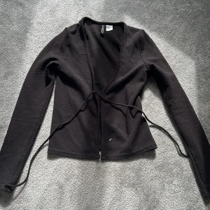 Svart omlottkofta från H&M Divided - Svart ribbad omlottkofta från H&M Divided i storlek XS. Koftan har långa ärmar och knytband framtill för en snygg passform. Materialet är mjukt och stickat, perfekt för lager-på-lager och chill stil. Enkel att matcha med jeans eller kjol.