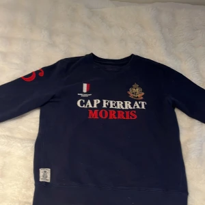 Mörkblå sweatshirt Morris Cap Ferrat - Snygg mörkblå sweatshirt från Morris Cap Ferrat i storlek medium. Tröjan har broderade detaljer med text och emblem på bröstet samt en stor röd siffra 6 på ena ärmen. Tillverkad i mjuk bomull med ribbade muddar vid hals, ärmslut och nederkant.