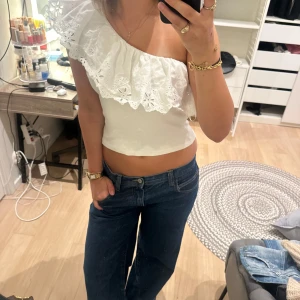 Vit omlott oneshoulder topp med volang - Superfin vit topp med bred volang och broderade spetsdetaljer. Sitter omlott över ena axeln och är croppad, perfekt till jeans eller kjol. Materialet känns som bomull och toppen har en oneshoulder-design som ger en cool och trendig vibe.