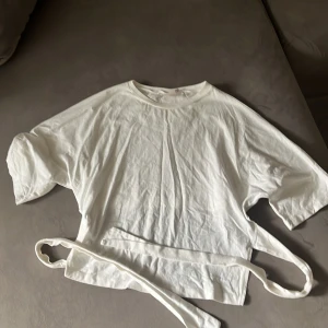 Vit omlottopp med knyt från Nelly - Vit omlottopp från Nelly i mjuk bomull. Modellen har korta ärmar, rund hals och långa knytband i midjan som ger en snygg siluett. Perfekt för dig som gillar minimalistisk stil med en twist. Passar dig som vill ha en enkel men trendig topp.