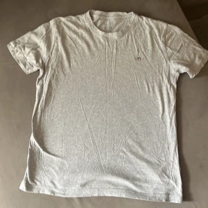 Grå t-shirt från GANT med logga - Snygg grå t-shirt från GANT med klassisk rund hals och korta ärmar. På bröstet sitter en liten broderad GANT-logga med sköld i rött, vitt och blått. Mjuk och skön bomull som passar perfekt till jeans eller shorts. Enkel och stilren design.