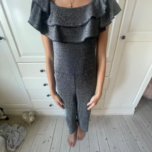 Glittrig jumpsuit från KappAhl - Säljer en snygg glittrig jumpsuit från KappAhl i storlek 134. Den är silverfärgad med croppade ben och har volangdetaljer upptill. Perfekt för dig som vill sticka ut med en cool och festlig look. Materialet är mjukt och stretchigt syntet.