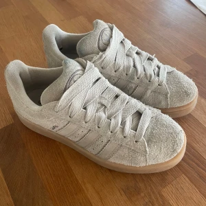 Adidas Campus sneakers i ljusbeige mocka - Adidas Campus sneakers i ljusbeige mocka med klassiska tre ränder på sidan och broderad logga på plösen. Skorna har grova snören och en platt gummisula i beige ton. Perfekt för dig som gillar retrovibe och clean stil. Inköpta i våras. 