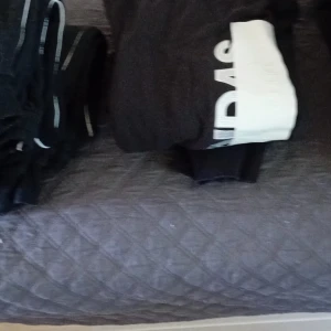 Svarta mjukisbyxor från Adidas - Svarta mjukisbyxor från Adidas med vit logga längs benet. Sköna och avslappnade med elastisk midja och ribbade muddar vid benslut. Perfekta för chill eller träning, tillverkade i mjukt bomullsmaterial.