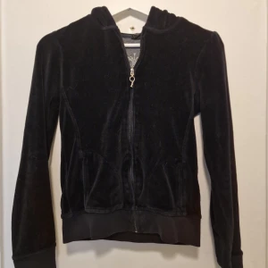 Svart mjuk velour hoodie - Svart hoodie i mjuk velour med silverfärgat kronmotiv och dekorativa detaljer på ryggen. Supermysig och lite mer åt lyxig streetstyle. En bit utav hänget på dragkedjan har gått av därav lågt pris. Se sista bilden.