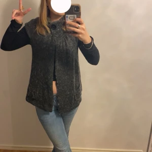 Mörkgrå stickad väst med knappar - Mysig mörkgrå stickad väst med två stora knappar vid halsen och lös passform. Västen är ärmlös och har en lätt melerad look, perfekt att slänga över en långärmad tröja. Enkel och stilren design som funkar till jeans eller kjol.