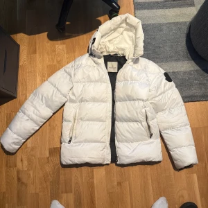 Vit Moncler puffer - Snygg vit dunjacka från Moncler med svart innerfoder och stor huva där Moncler-loggan är tryckt. Jackan har dragkedja framtill, två fickor med dragkedja och quiltad design. Perfekt för kalla dagar och ger en clean look.