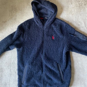 Polo Ralph Lauren tröja - Säljer nu denna Mysiga marinblå teddyjacka från Polo Ralph Lauren med huva och dragkedja. Jackan har en röd logga på bröstet, fickor framtill och en extra ficka med dragkedja på ärmen. Perfekt för kyliga dagar när du vill vara både varm och stilren.