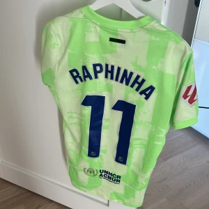 Barcelona Raphinha kortärmad matchtröja - Säljer en Barcelona fotbollströja med Raphinha och nummer 11 på ryggen. Tröjan är ljusgrön med ett unikt mönster och har klubbmärke, Nike-logga och Spotify som sponsor framtill. Materialet är lätt och andas, perfekt för träning eller match.