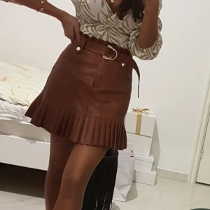 Brun skinnkjol med volang och bälte - Snygg brun kort skinnkjol med volangkant och tillhörande bälte i midjan. Kjolen har hög midja och markerad siluett, perfekt för dig som vill sticka ut. Stängs med knappar och har coola detaljer som ger en trendig look.