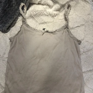 Beige ribbad linnetopp från Divided - Supersöt beige linnetopp från Divided med ribbad struktur och smala axelband dekorerade med spets. Toppen har en liten rosett framtill och är perfekt för att skapa en chill och stilren look. Materialet är mjukt och skönt bomull.