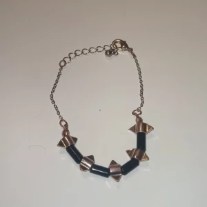 Svart och guldigt armband med nitar - Coolt armband med svarta stavar och guldiga pyramidformade nitar. Kedjan är tunn och har ett justerbart lås med en liten kula som detalj. Perfekt för dig som gillar edgy accessoarer med metalliska inslag.