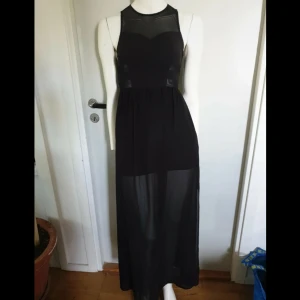 Svart långklänning med meshdetaljer - Snygg svart långklänning med transparent mesh upptill och i midjan. Klänningen har ärmlös design, rund hals och dragkedja i ryggen. Kjolen är lätt och har en skir effekt nedtill. Perfekt för dig som gillar en stilren look med coola detaljer.