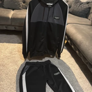 Svart och grå Tracksuit från McKenzie - Snygg svart och grå Tracksuit från McKenzie med huva och dragkedja framtill. Jackan har vita ränder längs ärmarna och ett stilrent McKenzie-märke på bröstet. Materialet är mjukt och syntetiskt, perfekt för chill och sportiga dagar. Långärmad och loose fit.