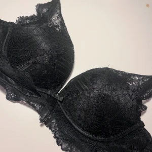 Svart pushup BH med spets - Supersnygg svart push up BH med spetsdetaljer och liten rosett framtill. BHn har push up kupor och tunna axelband, samt vackert mönstrad spets längs kanterna. I mycket bra skick